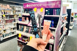 person holding essence mascara, eyeliner, and lip gloss in front of a makeup display