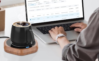 a coffee mug on a mug warmer near a person on a laptop 