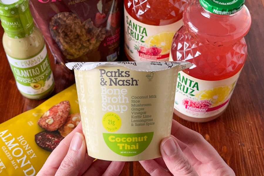 A person's hands holding a container of Parkins & Nash bone broth soup above other organic products sitting on a wooden table.