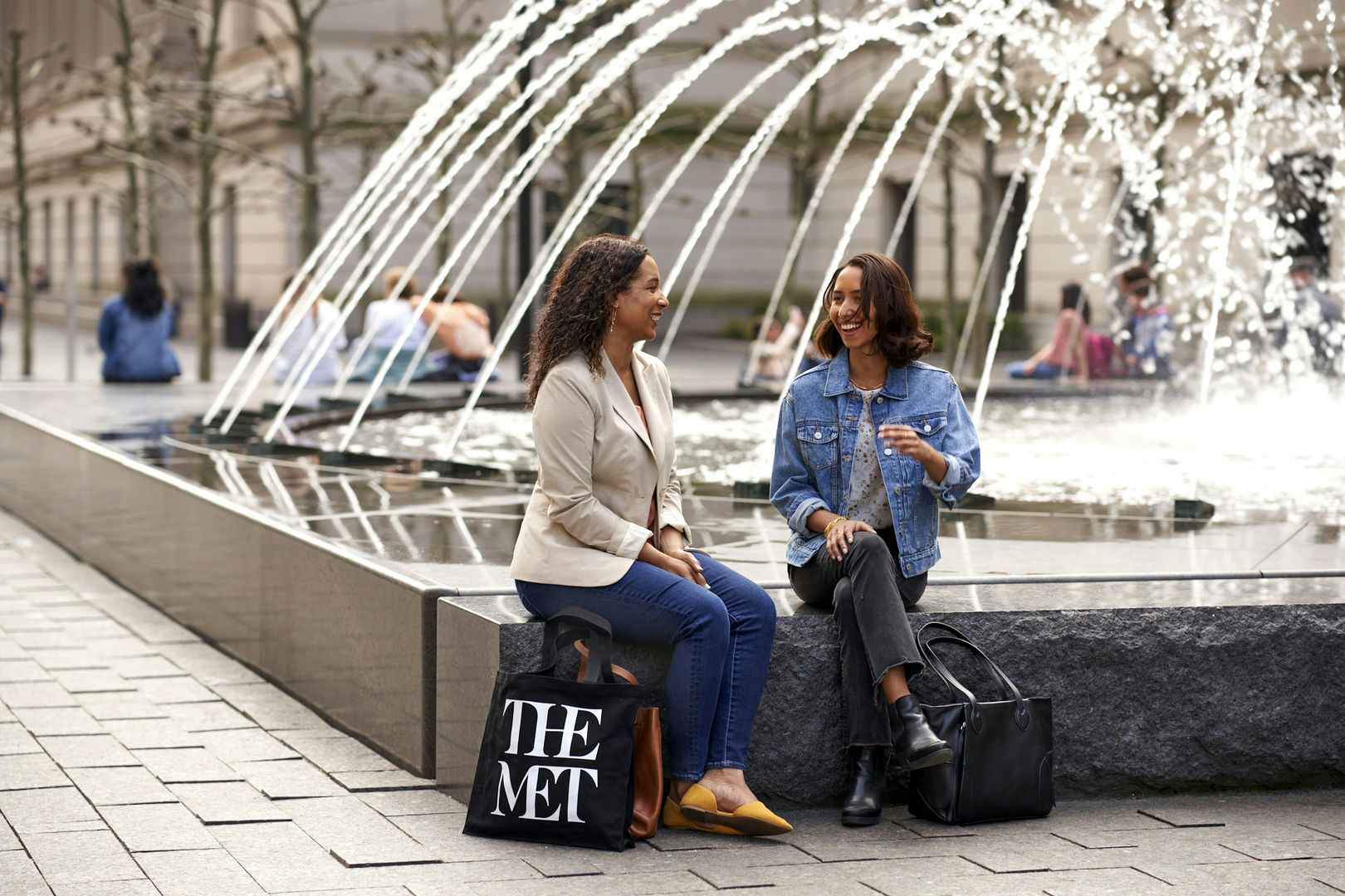 Two people having a conversation while sitting on a fountain outside of The Metropolitan Museum of Art with bags that say "The Met"