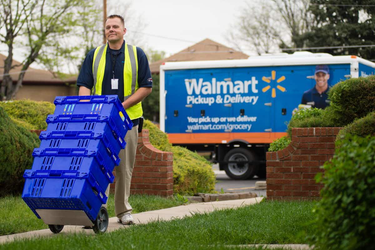 walmart worker wheels groceries up home sidewalk with walmart delivery truck in background