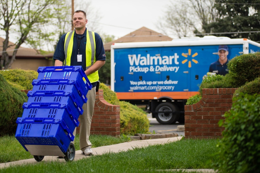 walmart worker wheels groceries up home sidewalk with walmart delivery truck in background