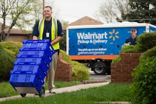 walmart worker wheels groceries up home sidewalk with walmart delivery truck in background
