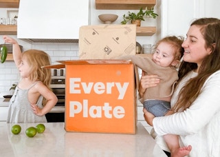 A mom holding her child as the child reaches for an Every Plate box sitting on the kitchen counter and another child holding up a ...