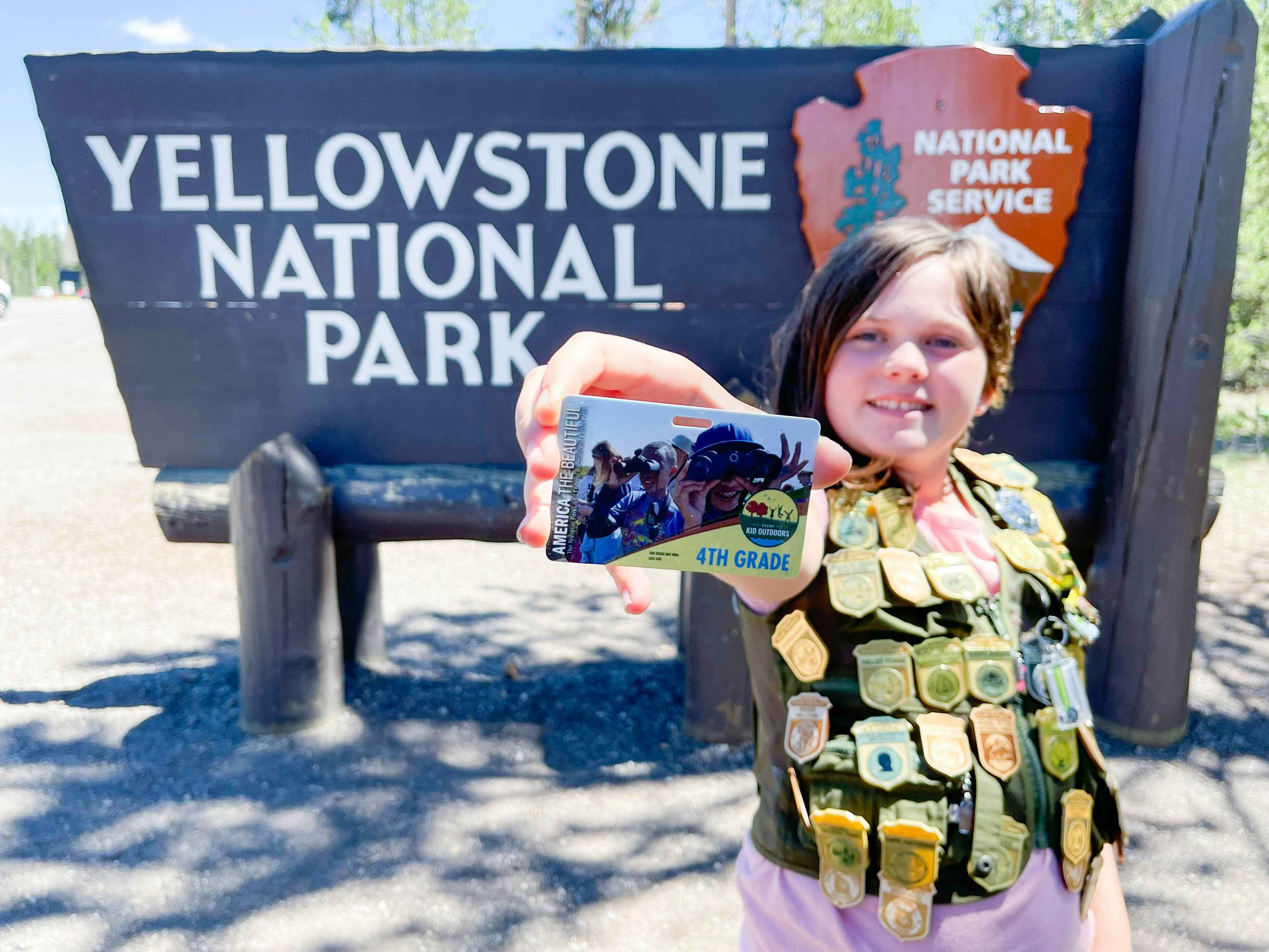 A child in front of the yellow stone national park sign wearing a vest covered in junior ranger badges. The child is holding out a free e...