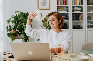 A woman smiles at her computer and holds cash in the air