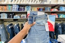 someone holding up a pair of jeans in front of multiple jeans hanging up, and t-shirts on a shelf in old navy