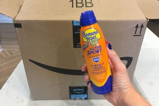 A hand holding a bottle of Banana Boat sunscreen in front of a delivery box.