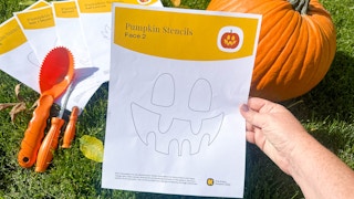a pumpkin stencil template with a face being help in front of a pumpkin and tools sitting in the grass