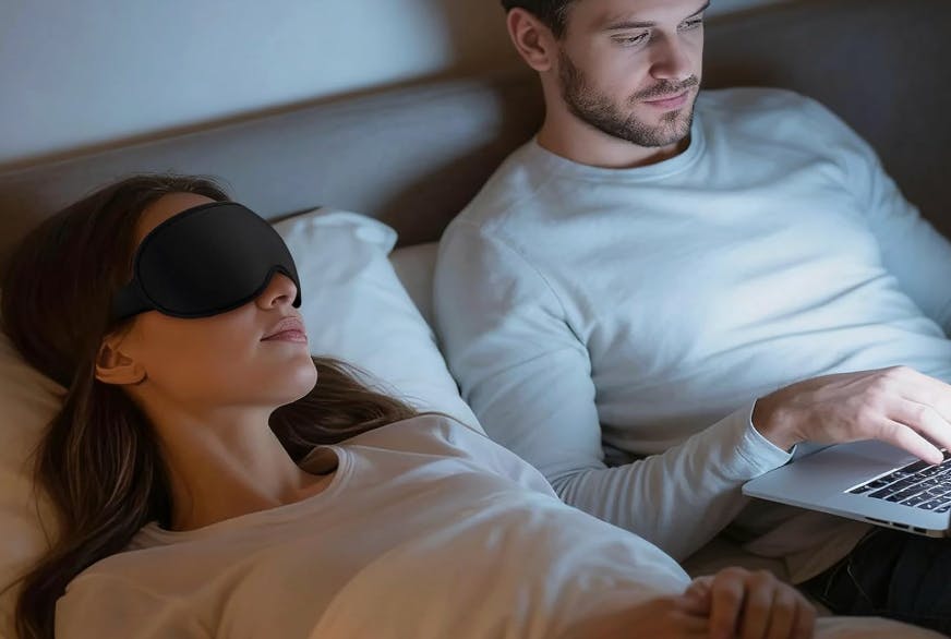 a person laying with a sleep mask next to a person on a laptop