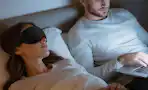 a person laying with a sleep mask next to a person on a laptop