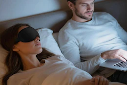 a person laying with a sleep mask next to a person on a laptop