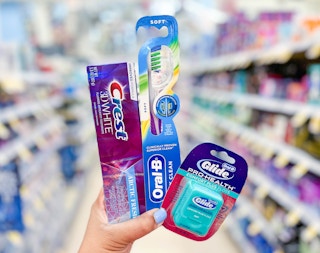 Hand holding one Crest 3D White toothpaste, one Oral-B Manual Toothbrush, and one Oral-B Glide Pro-Health Floss in aisle