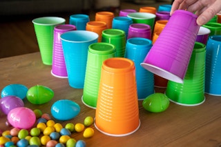 Pink, green, orange, and blue plastic cups set out on a table in a pattern with plastic Easter eggs under them and next to them.