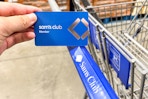 a person holding up a sams club card in front of a sams club cart
