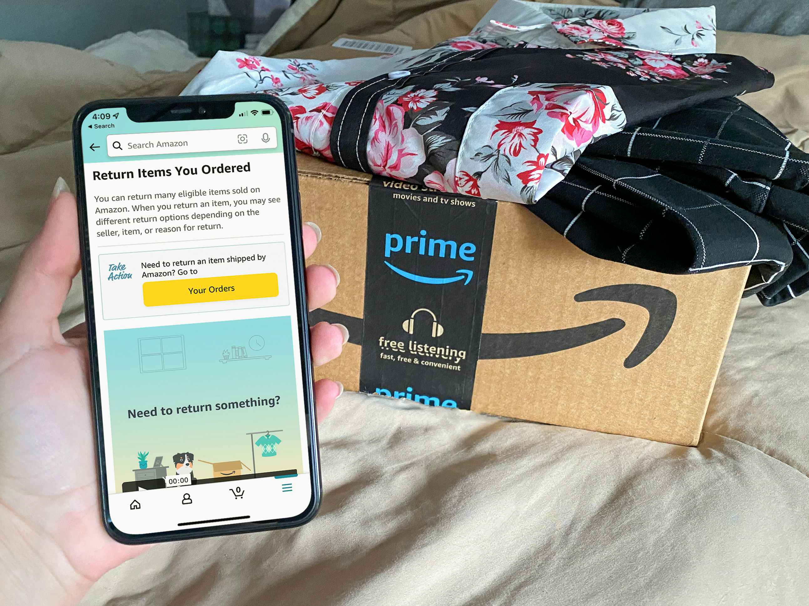 A person's hand holding an iPhone displaying the "Return Items You Ordered" page on the Amazon app next to an Amazon Prime box with cloth...