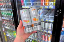 person holding 2 celsius energy drinks in front of a cooler