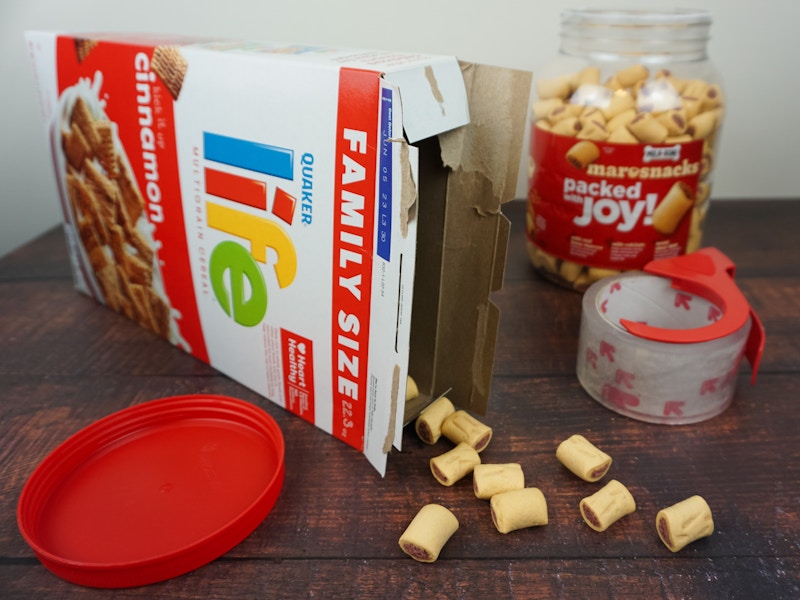 A cereal box with treats coming out of it on a table next to a plastic tub of dog treats and a roll of packing tape