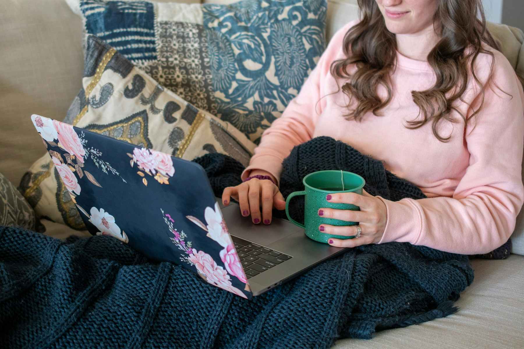 A woman sitting on a couch, looking at a laptop with a cup of tea in her hand.