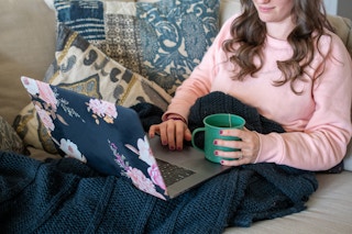 A woman sitting on a couch, looking at a laptop with a cup of tea in her hand.