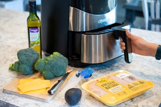 A person opening an air fryer with chicken, cheese, broccoli, olive oil, and an avocado nearby.