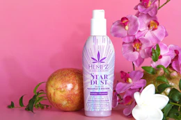 A bottle of Hempz Star Dust Body Oil surrounded by flowers and fruit