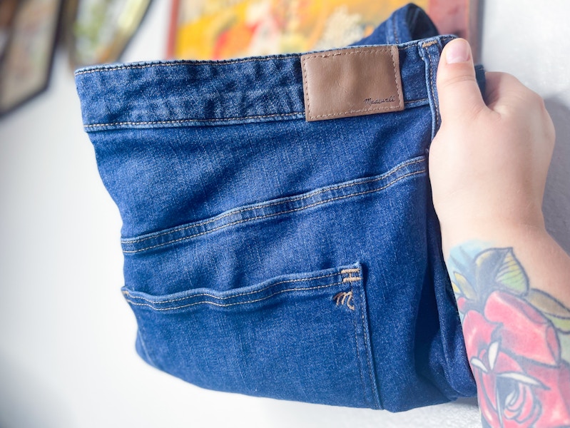 A person's hand holding up a pair of Madewell jeans in their home.