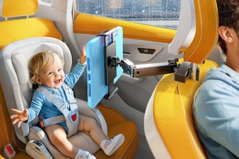 A baby sits in the back seat in a carseat, looking happily at a tablet on the arm of the tablet holder, which is attached to the headrest