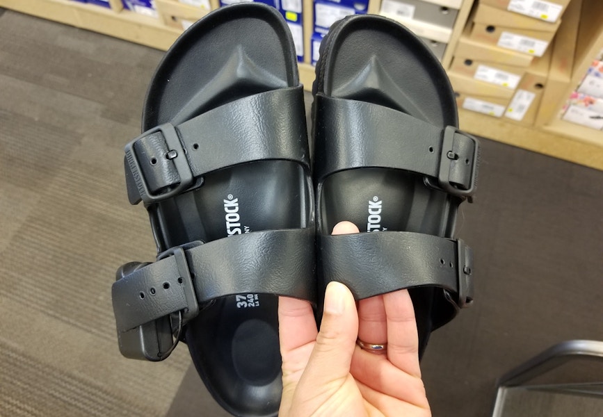 A person's hand holding up a pair of Birkenstock sandals in a shoe store.