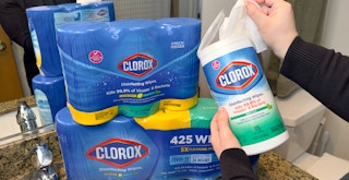Someone taking a wipe out of a Clorox container next to a stack of multi-packs of disinfectant wipes on a bathroom counter
