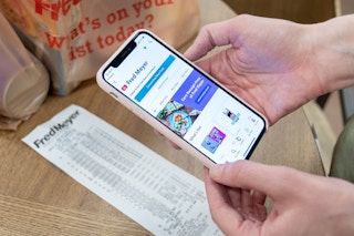 A person's hands holding an iPhone with the Ibotta mobile app open about to scan a receipt that is on a table next to two bags of groceri...