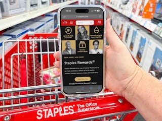 A person's hand resting on the handle of a Staples shopping cart, holding a cell phone displaying the Staples Rewards page on their website.