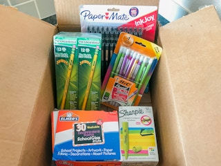 A box filled with school supplies including pencils, mechanical pencils, and glue sticks.