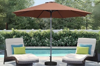 A brown 9-foot patio umbrella stands between two white lounge chairs with blue and green pillows