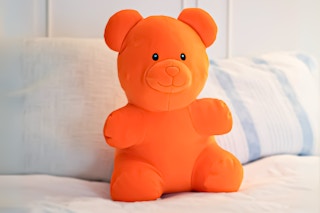 Lifestyle image of the orange Way to Celebrate Gummy Bear Plush on bed