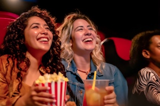 two women watching a movie and holding a drink and popcorn 