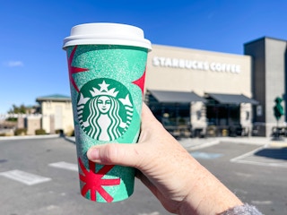 a person's hand holding up a starbucks holiday drink outside starbucks store front