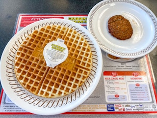 A plate with a big waffle and a container of Waffle House butter sitting next to another plate with a sausage patty on a table at Waffle ...