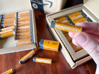 A person holding up an orange amazon basics battery with an open box of batteries in the background.