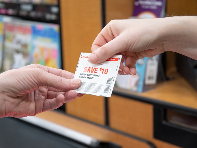 Two people's hands, one handing the other a coupon for $10 off at the checkout lane in a store.