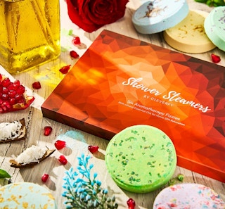 Red and orange box on a wooden table surrounded by shower steamers, herbs,and an oil bottle