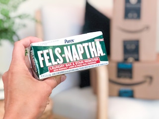 A hand holding up a Purex Fels-Naptha laundry bar in front of Amazon boxes.