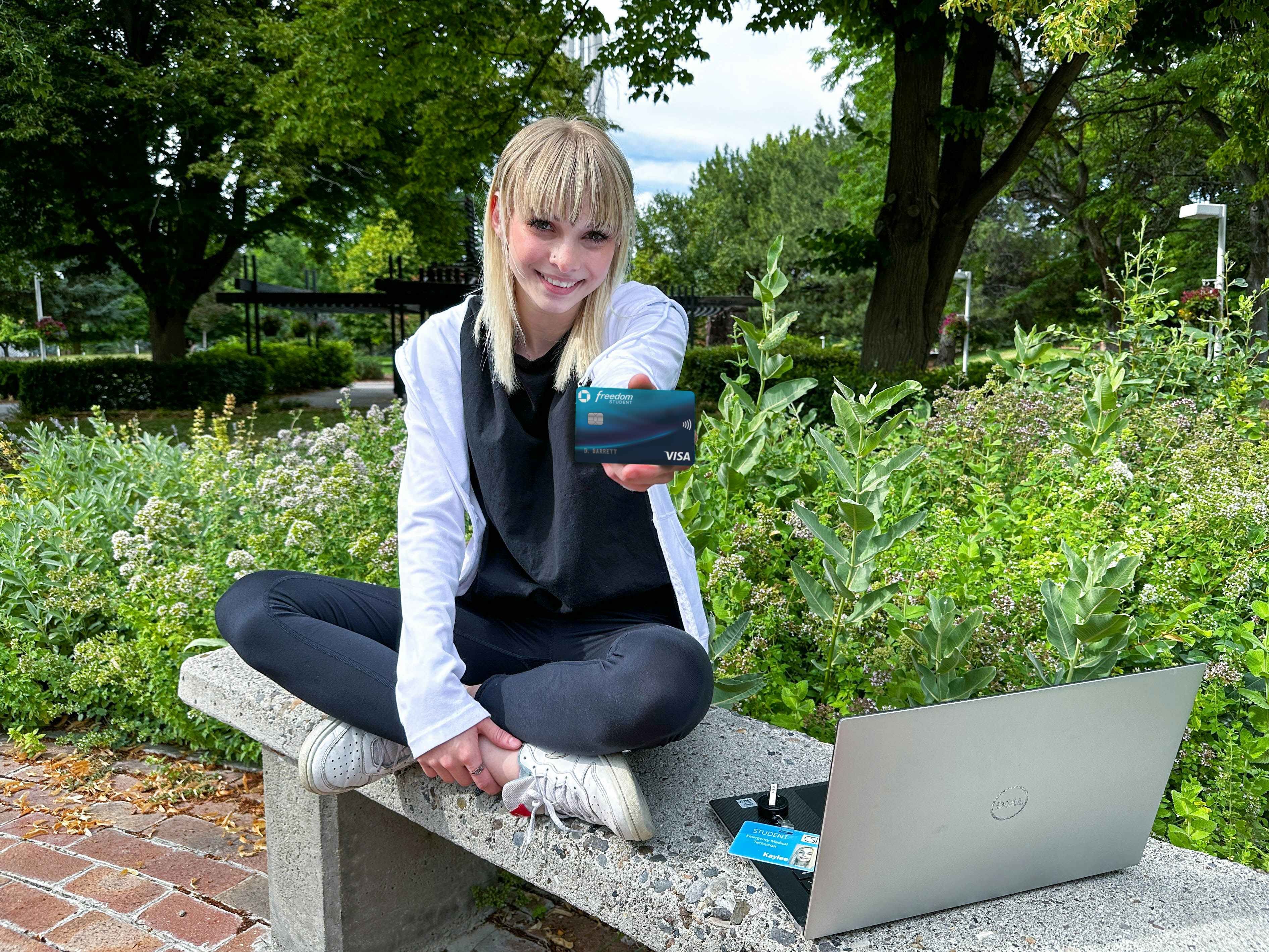 a college student sitting on a bench outside on campus with laptop holding out a debit credit card 