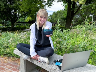 a college student sitting on a bench outside on campus with laptop holding out a debit credit card 