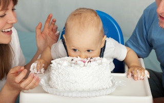 A baby sticking their face into a cake with the parents on either side, smiling