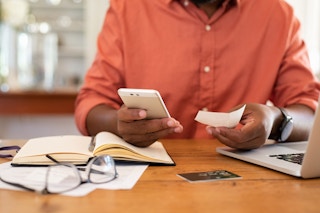 A person holding a phone and looking at a bill with a credit card and documents on the table