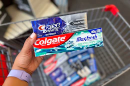 hand holding tubes of crest and colgate toothpaste over a shopping cart full of toothpaste