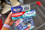 hand holding tubes of crest and colgate toothpaste over a shopping cart full of toothpaste
