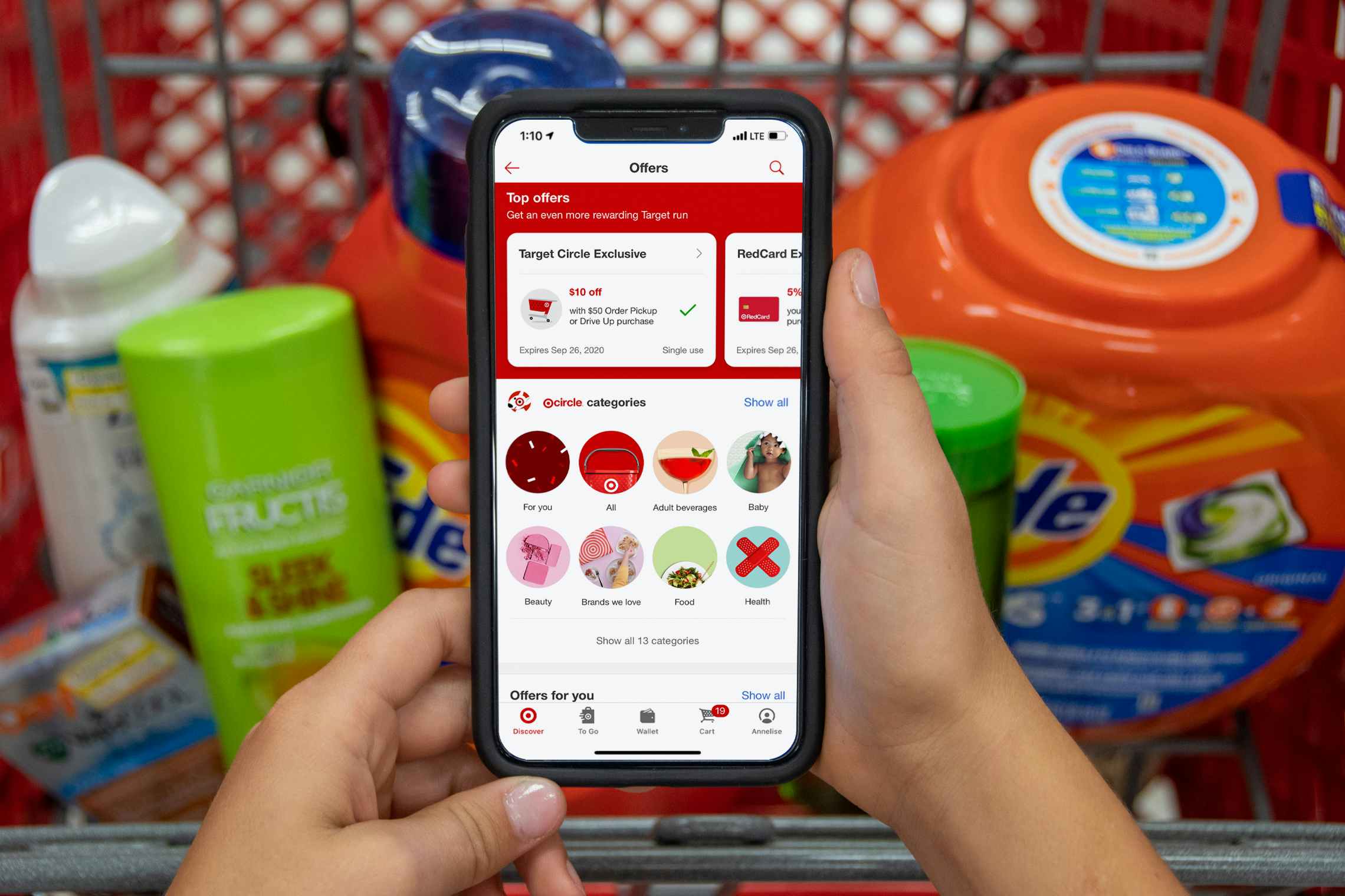 A person's hands holding a phone displaying the Target mobile app's Offers page in front of a Target shopping cart's basket filled with ...