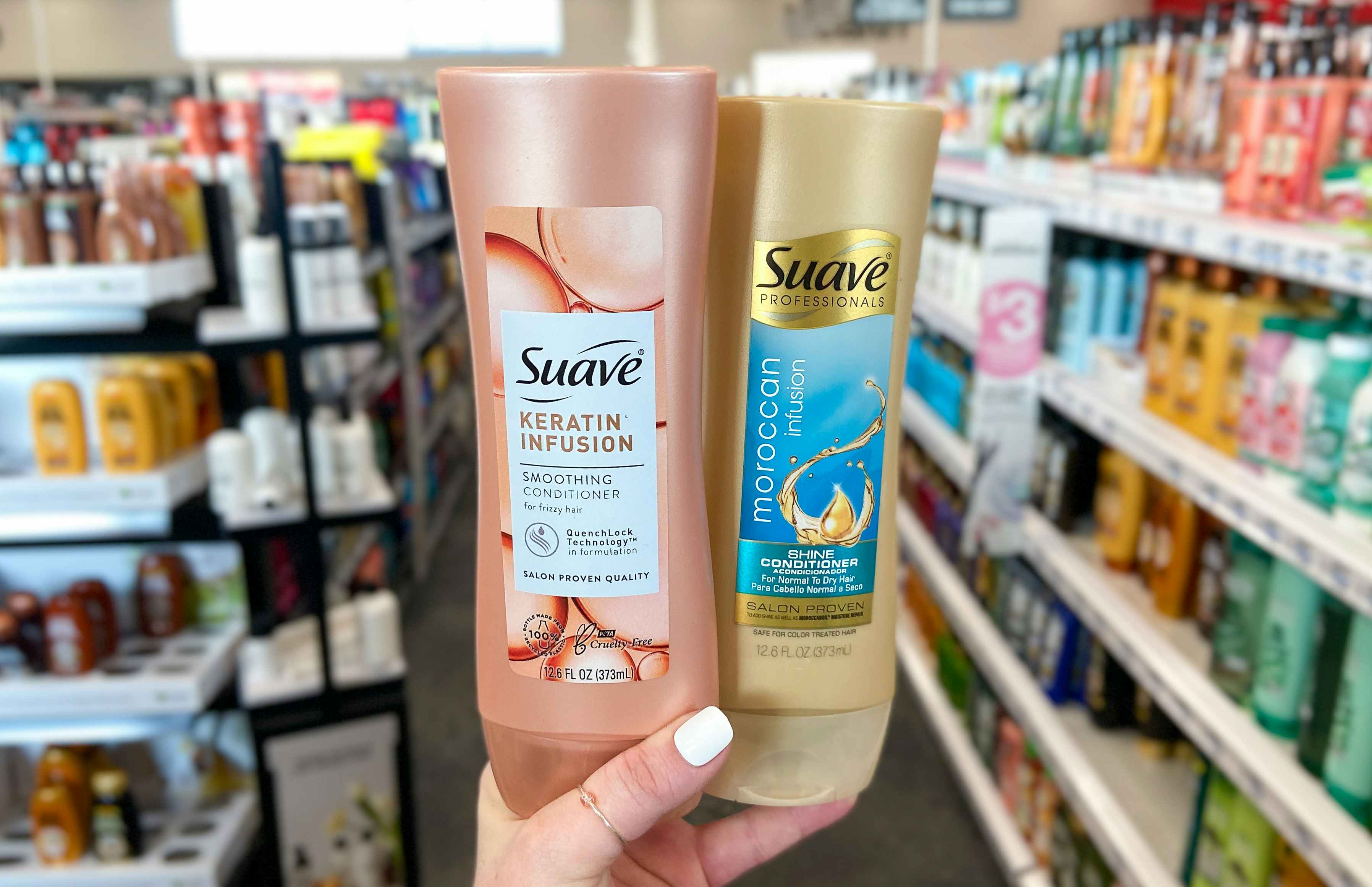 A person's hand holding up two bottles of Suave conditioner in front of an aisle in a store. One is Suave Keratin Infusion smoothing cond...